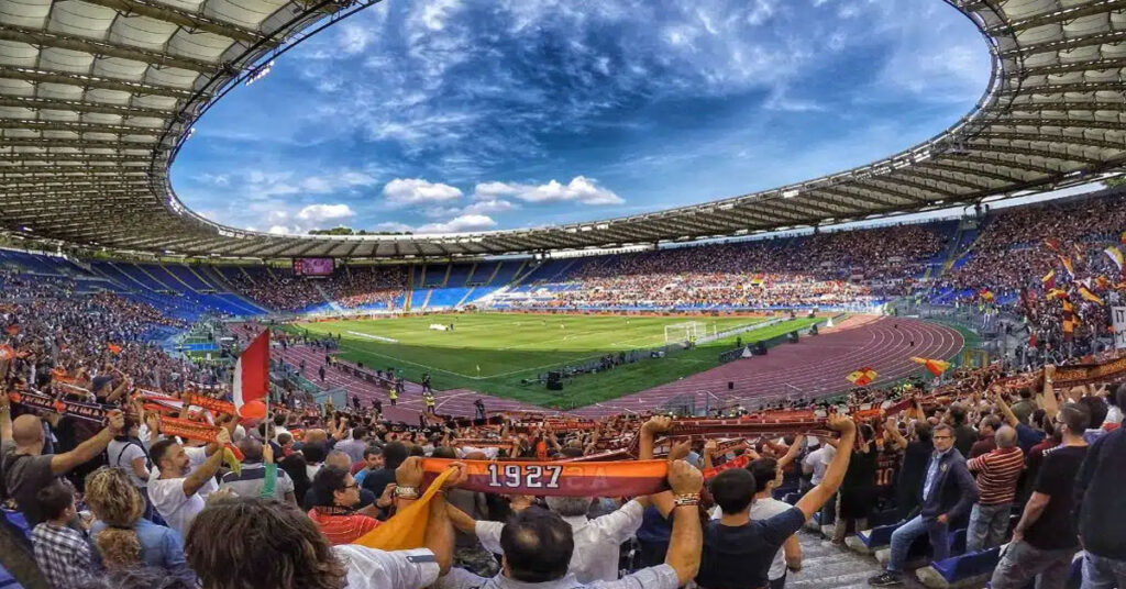 Large football stadium filled with spectators during a daytime match under a bright but partly cloudy sky.