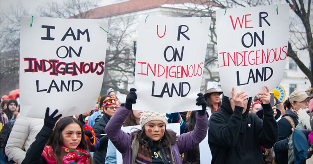 Demonstrators holding signs reading “I am on Indigenous Land” during a public protest advocating land rights.