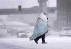 A person wrapped in blankets crosses a snow-covered road while vehicles drive slowly through low visibility during a winter storm.