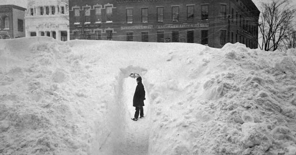A person stands inside a narrow passage carved through towering snowbanks in a city buried under heavy snowfall.