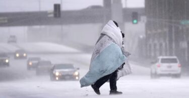A person wrapped in blankets crosses a snow-covered road while vehicles drive slowly through low visibility during a winter storm.