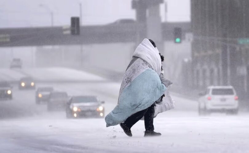 A person wrapped in blankets crosses a snow-covered road while vehicles drive slowly through low visibility during a winter storm.