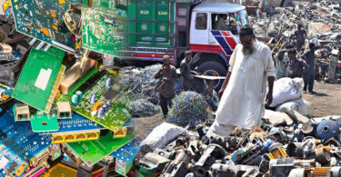 Workers sorting electronic scrap and wires in an open scrapyard surrounded by piles of discarded equipment and debris.