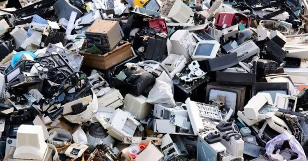 Workers sorting electronic scrap and wires in an open scrapyard surrounded by piles of discarded equipment and debris.